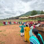 Children in bright clothes form a circle on a dirt field with uniformed officials standing along the left and tents with chairs on the right under a canopy